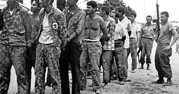 Members of 2506 Brigade march in a Cuban prision camp guarded by armed men.
